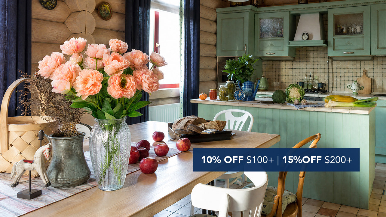 Faux pink peony floral arrangement in a glass vase on a rustic cottage dining table with 10% OFF $100+ and 15% OFF $200+ promotional banner.