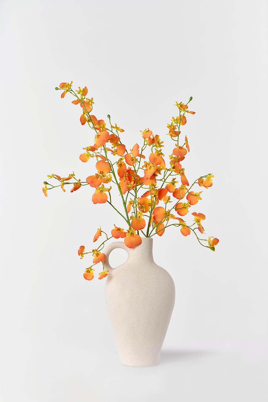 A white vase filled with artificial dancing orchid flowers in shades of orange. The blooms look like they are in full bloom, adding a lively feel to the arrangement.