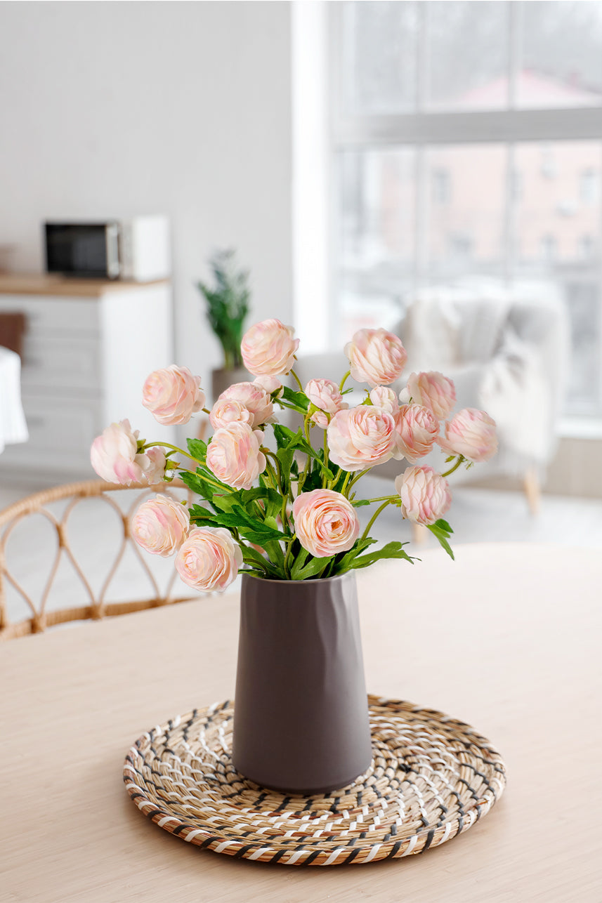 The artificial light pink ranunculus and the light grey vase together create a perfect floral arrangement which is placed on the dining table.