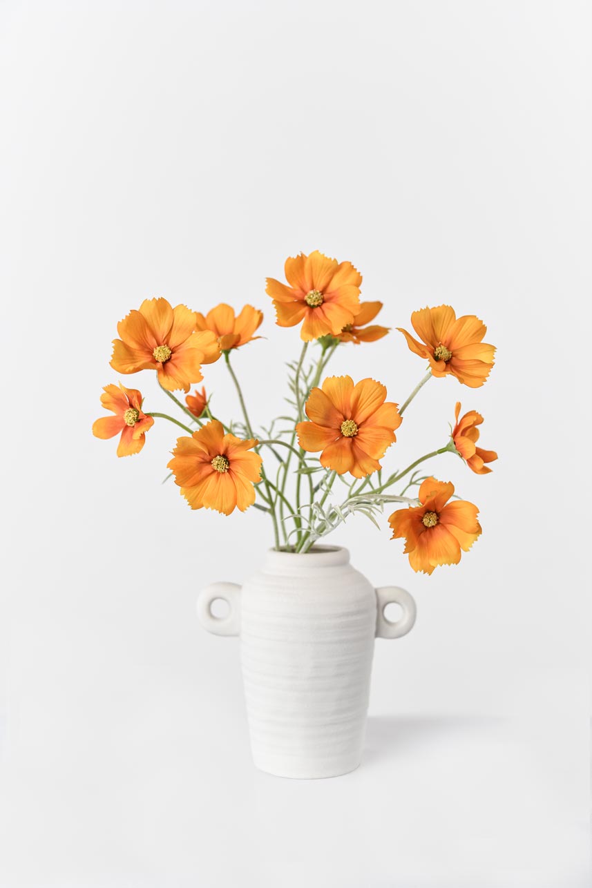 Realistic faux orange Cosmos bipinnatus flowers arranged in a white vase, adding a touch of natural beauty to your decor.