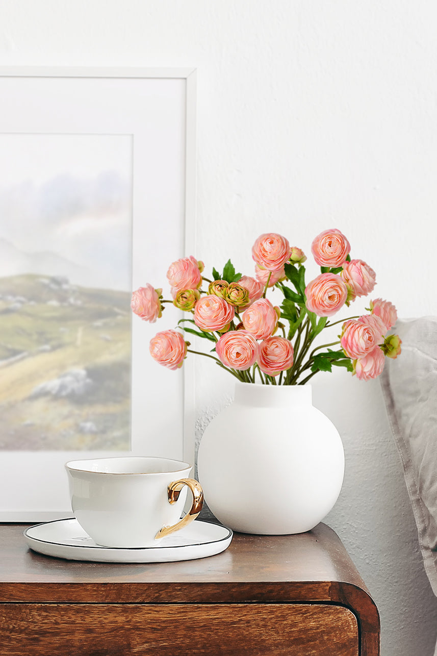 Captivating pink Ranunculus artificial flowers grace a white vase, enhancing the coffee table with their timeless beauty.