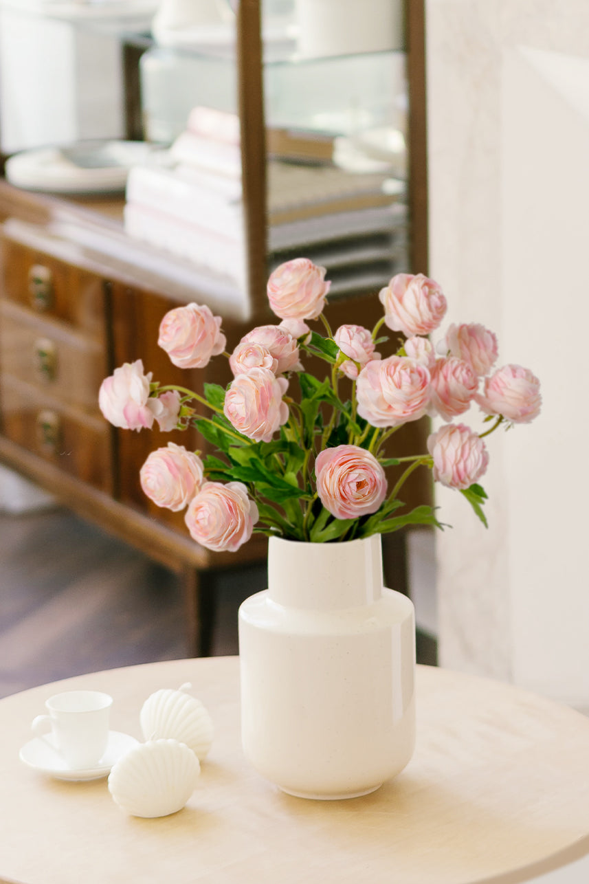 Soft and delicate, light pink Ranunculus silk flowers create a charming display in a white vase, adding a touch of elegance to any tabletop.