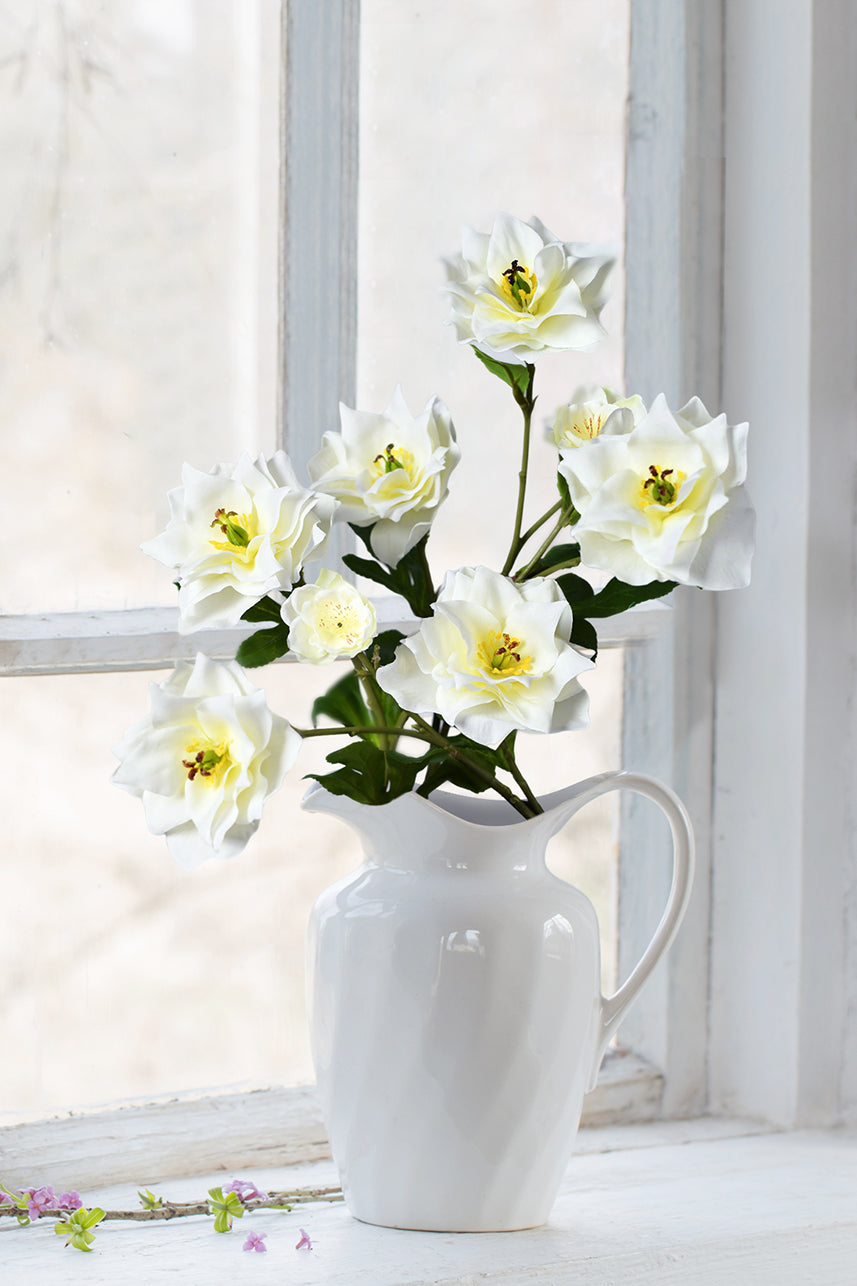 Cream-white Clematis faux blooms adorn a white vase, beautifully positioned near a window for a touch of natural grace.
