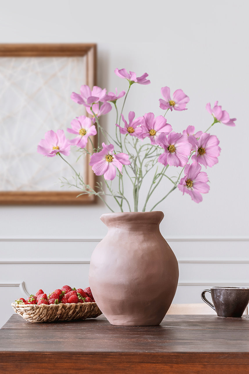 Lavender pink artificial cosmos bipinnatus are placed in a brown ceramic vase with strawberries and teacups around the vase.